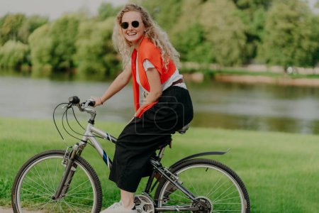 Photo for Pleasant looking satisfied curly haired woman spends free time in open air, rides bicycle, spends leisure alone, enjoys freshness of nature, looks at camera with pleasant smile. Summer time. - Royalty Free Image
