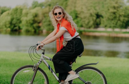 Photo for Pretty active female bicyclist rides bike, looks at camera with smile, wears casual everyday clothes, poses near river, green trees, grass, spends free time in countryside. People, rest, lifestyle - Royalty Free Image