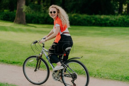 Photo for Outdoor image of happy woman rides bike on path, green park around, wears sunglasses, t shirt, pants and sportshoes, has summer holiday, spends spare time actively. People, healthy lifestyle - Royalty Free Image