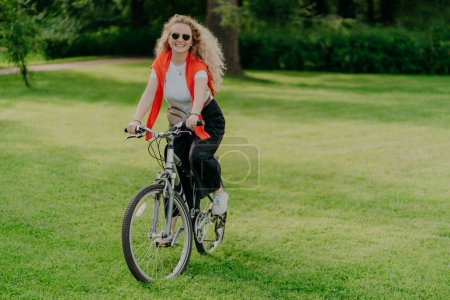 Photo for Positive smiling woman dressed in casual clothes, poses on bicycle, has active rest in park, rides on green lawn during sunny summer day, being in high spirit. People, recreation, hobby concept - Royalty Free Image