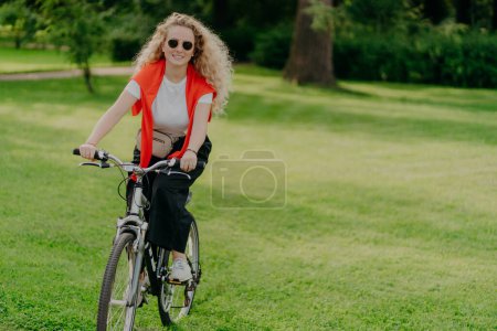 Photo for People, nature, rest, lifestyle concept. Happy curly woman rides bicycle among green grass, moves actively, wants to be fit, explores new places in countryside, wears sunglasses, casual clothes - Royalty Free Image