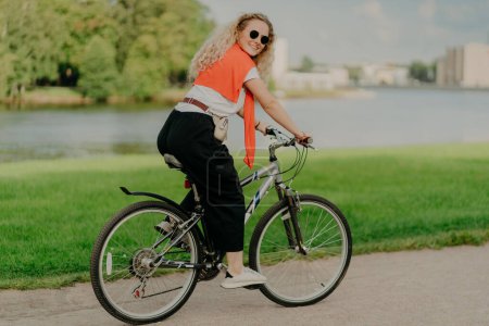 Photo for Pleased young woman enjoys new route on bicycle, rides among lake, green lawn and buildings far away into distance, wears summer shades, casual outfit, white sneakers, being in good physical shape - Royalty Free Image