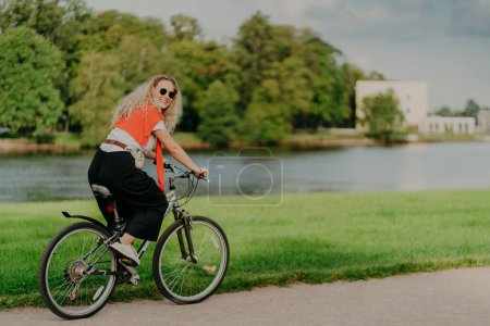 Photo for Photo of cheerful woman dressed casually, rides bicycle, looks aside, has happy expression, wears shades, poses near river, green lawn and trees, some buildings in background. People and rest - Royalty Free Image