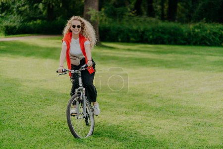 Photo for Outdoor image of pretty cheerful young woman rides bike, wears sunglasses, casual wear, poses on green lawn, spends free time in park, bikes in beautiful nature. Activity and recreation concept - Royalty Free Image
