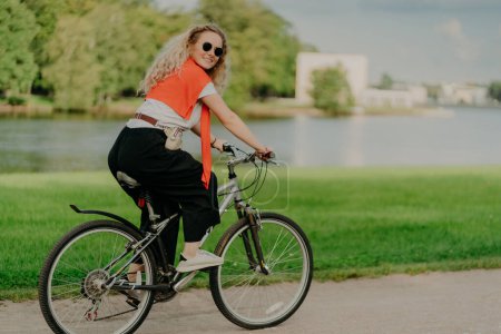 Photo for Image of female model rides bicycle, looks aside with cheerful expression, wears sunglasses, breathes fresh air, poses near lake and green trees, covers long distance, finds out something new - Royalty Free Image