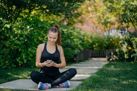 Photo for Outdoor shot of athletic woman rests after workout sits crossed legs in park surfs social networks dressed in sportswear listens music via wireless earphones uses sport app enjoys good weather - Royalty Free Image