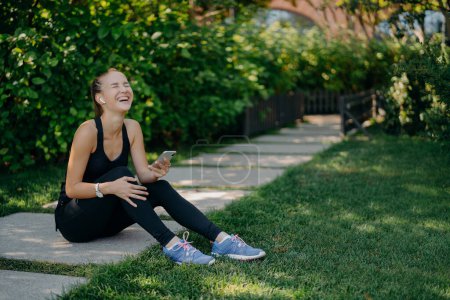 Photo for Overjoyed sportswoman sits on path laughs out uses smartphone for listening music from playlist share content and chatting dressed in active wear enjoys spare time. Active lifestyle concept. - Royalty Free Image