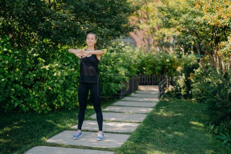 Photo for Full length shot of active fit motivated woman warms up hands before training dressed in black sportsclothes and sneakers does physical outdoors in open air breathes deeply leads healthy lifestyle. - Royalty Free Image