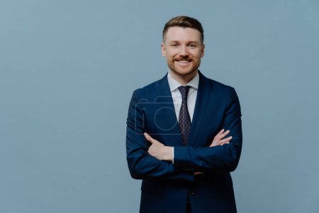 Photo for Waist up shot of self confident bearded male entrepreneur keeps arms crossed smiles gladfully dressed in formal clothes isolated over blue studio background copy space for your advertising content - Royalty Free Image