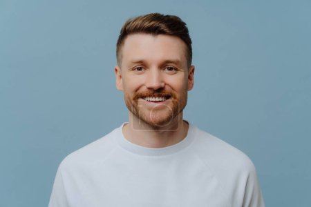 Headpshot portrait of young happy unshaven man in white tshirt looking at camera with excited facial expresson while posing against blue studio background. Human emotions and positivity concept