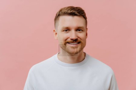 Portrait of handsome pleasant unshaven redhead man expressing positiveness while looking at camera with smile, happy guy in white tshirt feeling joyful while posing against pink studio background.