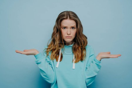 Photo for Confused young female in casual hoodie cannot make decision, spreading arms with facial expression of doubt and hesitation, sad teenage girl expressing uncertainty while posing against blue background - Royalty Free Image