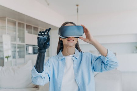 Photo for Disabled person gets rehabilitation. Handicapped girl with cyber arm in vr goggles at home. Young european woman sitting in living room on couch is excited. Lady touches the vision. - Royalty Free Image