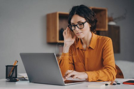Freelance. Beautiful focused Spanish woman in glasses sits at desk in living room types communicates with client live on laptop, focused young female consultant adviser working remotely online at home