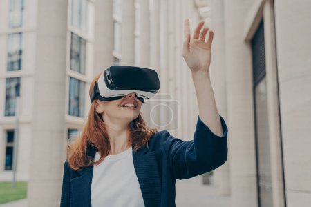 Photo for Redhead businesswoman managing business project through virtual reality platform. Smiling young female office worker enjoying spending time with VR headset glasses, pointing with forefinger up in air - Royalty Free Image