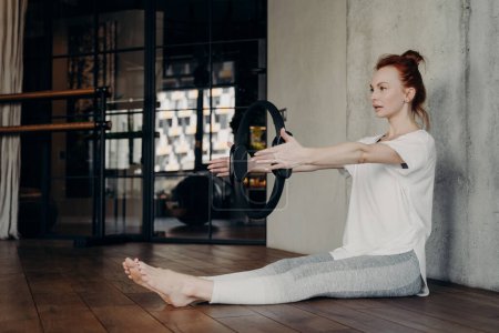 Seitenansicht einer jungen, fokussierten, sportlichen Frau, die mit ausgestreckten Beinen auf dem Boden im Fitnessstudio sitzt und mit Pilatesring Armübungen macht, weißes Fitness-T-Shirt und graue Leggings trägt