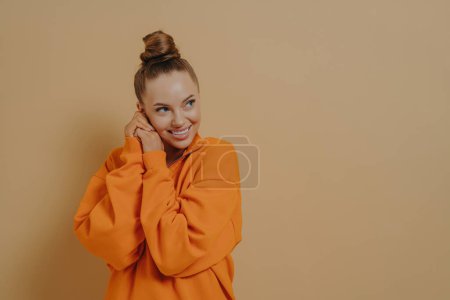 Photo for Young charming shy woman flirting and showing attraction, pressing hands together near face and smiling, happy to hear compliment, dressed in orange sweatshirt standing isolated over beige background - Royalty Free Image
