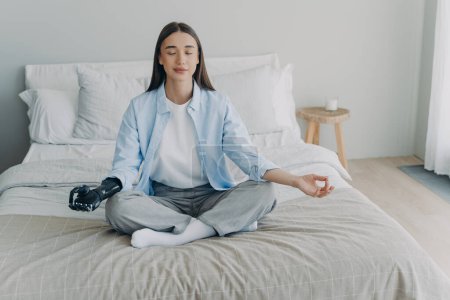 Young disabled woman is sitting in lotus pose on bed with her eyes closed. Attractive european girl has cyber prosthesis. Modern artificial limb. Handicapped woman lifestyle concept.