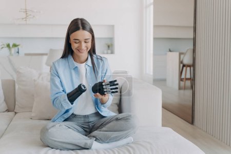 Happy disabled young woman is disassembling bionic arm prosthesis. Female amputee adjusting her artificial arm at home. European girl has myoelectric carbon hand. Electronic settings.