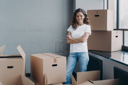 Foto de Satisfied woman unpacking boxes. Attractive girl in new apartment. Young woman in jeans and white t-shirt. Relocation, delivery service and new apartment concept. - Imagen libre de derechos