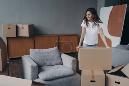 Photo for Smiling european woman is packing things. Girl moves to new house. Young lady is unpacking cardboard boxes in new apartment. Big new living room with armchair. Real estate purchase concept. - Royalty Free Image