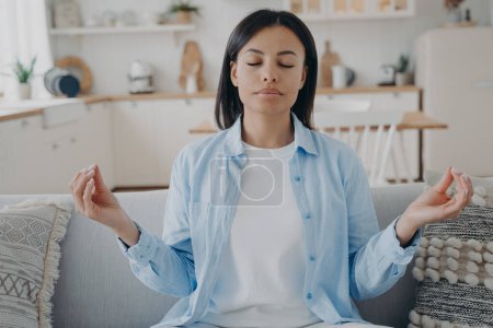 Photo for Calm woman meditate folded fingers in mudra hand gesture sitting on sofa at home. Peaceful young female practice yoga, relieve negative emotions, relax on couch in living room. Emotion management. - Royalty Free Image