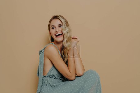 Téléchargez les photos : Femme blonde joyeuse et insouciante en robe d'été romantique longue riant et exprimant le bonheur, belle femme regardant de côté et s'amusant tout en posant sur fond beige en studio - en image libre de droit