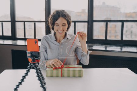 Photo for Excited hispanic woman blogger open gift box recording indoor video with xmas present for her blog on smartphone, female influencer sitting in front of mobile phone on stabilizer and unpucking present - Royalty Free Image
