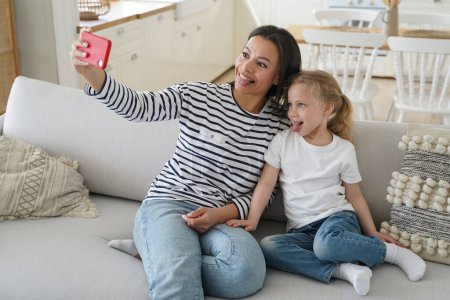 Téléchargez les photos : Funny little daughter and young mother showing tongue taking pictures of family, looking at mobile phone camera, happy mom having fun with kid girl, taking selfie together, sitting on couch at home. - en image libre de droit