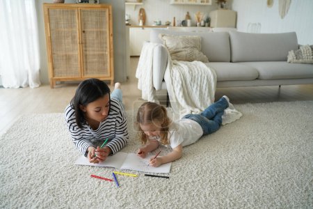Mother or babysitter and little kid girl lying on floor carpet in living room drawing together, mom with daughter painting enjoy creative hobby. Child creativity development, parenting concept.