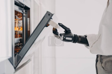 Female amputee is cooking using futuristic high technology bionic prosthesis. Robotic cyber hand of disabled person, which is standing in front of oven at kitchen. Daily routine of disabled.