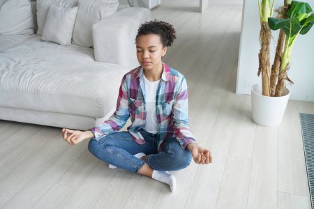Photo for Young biracial girl doing yoga sitting in padmasana, lotus pose on floor indoors. African american woman meditating, doing breathing exercises at home Interior. Stress relief, wellness concept. - Royalty Free Image