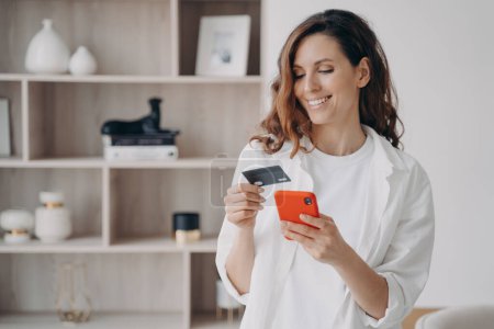 Foto de Easy payment with smartphone and card. Happy caucasian girl is going to buy through internet at home. Girl is booking or purchasing online. E-commerce and financial transaction concept. - Imagen libre de derechos