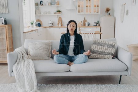 Photo for Serene woman practice yoga with mudra gesture, breathing sitting on sofa in living room. Young female meditate relieve stress, relaxing at home. Healthy lifestyle, emotion management. - Royalty Free Image