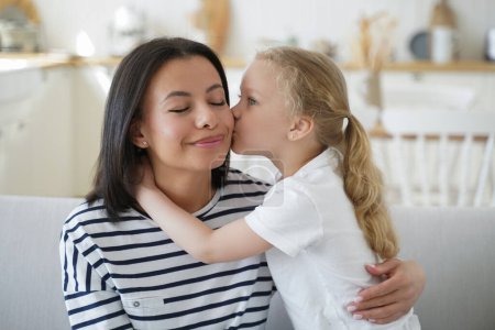 Kleine Mädchen Ziehtochter küssen Kuschelmutter zeigt Liebe zu Hause. Die lächelnde Mutter genießt zusammen mit ihrem Adoptivkind den netten, zärtlichen Moment. Glückliche Mutterschaft, Adoption von Kindern.