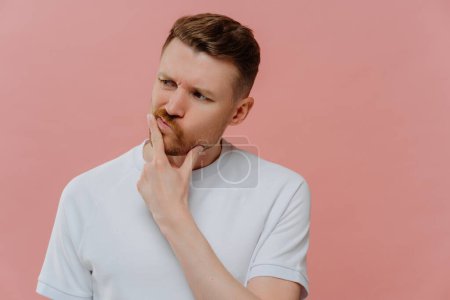 Photo for Thoughtful young unshaven man in white shirt having doubts and thinking about something with confused face expression while standing isolated over pink background. Hesitation concept - Royalty Free Image