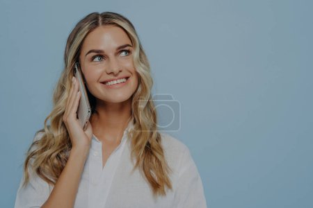 Photo for Joyful light headed female talking on phone with her friend, chatting about different things and sharing stories with each other, smiling while standing against blue background with copy space - Royalty Free Image