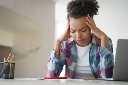 Tired african american teenage schoolgirl suffer headache while doing homework at laptop at home. Exhausted, frustrated teenager girl worried about educational problem or bullying. Difficult elearning