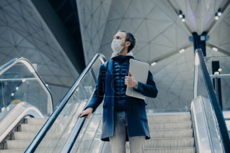 Photo for Shot of man tourist poses on escalator in airport, arrives home from abroad during virus outbreak, wears protective medical mask. Evacuated passenger. Infectious disease. Public safety concept - Royalty Free Image