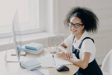 Positive Afro American female freelancer poses at workplace with papers and textbooks, works remotely on computer, has coffee break, works in her own cabinet, wears casual clothes. People, job Poster 647901914