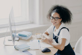Positive Afro American female freelancer poses at workplace with papers and textbooks, works remotely on computer, has coffee break, works in her own cabinet, wears casual clothes. People, job Poster #647901914