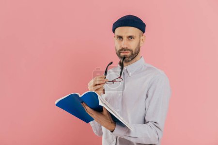 Photo for Self confident serious man with dark stubble, takes off spectacles, has attentive look at camera, holds opened textbook, learns necessary information, poses against pink background. Education - Royalty Free Image