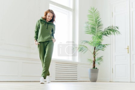 Photo for Full length shot of active pleased young woman with slim body, keeps hands in pockets of tracksuit, focused down, ready for doing cardio exercises, has determined look, pilates exercising indoor - Royalty Free Image