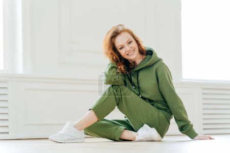 Photo for Delighted happy foxy woman busy with stretching pilate exercises in fitness studio, wears sportswear, has positive smile on face, enjoys workout, does cardio training. Acrobatics and sport concept - Royalty Free Image