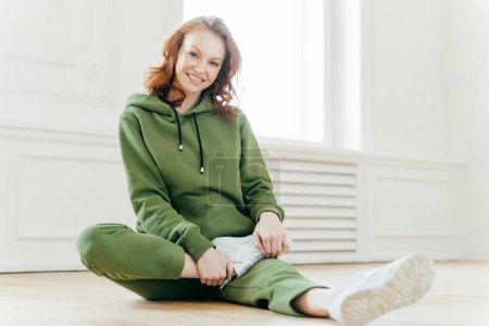 Photo for Photo of happy red haired woman in sweatsuit, white sneakers, takes rest after workout, sits on floor, stretches legs, has active healthy lifestyle. Smiling fitness girl does exercising. Flexibility - Royalty Free Image