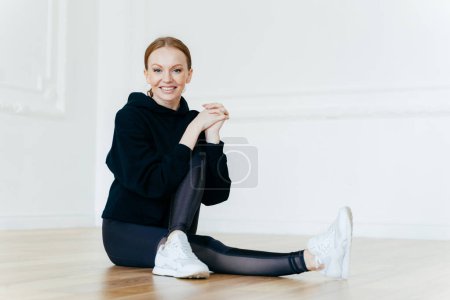Photo for People, sport, recreation concept. Delighted red haired female has positive smile, sits on floor in empty room, leads healthy lifestyle, does fitness exercises, wears hoody, leggings and sneakers - Royalty Free Image