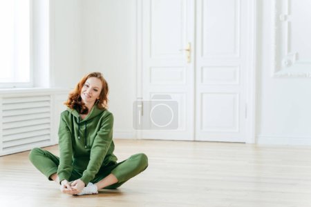 Photo for Photo of delighted redhead female sits on lotus pose, wears tracksuit, being in good body shape, has curly ginger hair, poses on floor, copy space for your promotional content. Sport concept - Royalty Free Image