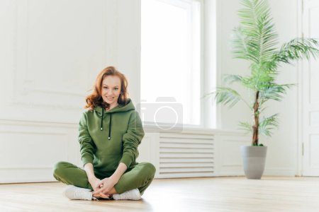 Photo for Beautiful satisfied slim woman with ginger hair, appealing appearance, sits crossed legs, wears green sweatsuit, keeps perfect body shape, stays fit. Athletic training and healthy lifestyle concept - Royalty Free Image