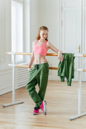Photo for Beautiful fit ginger female dancer has rest near barre in ballet dancing studio, focused down with thoughtful expression, wears top, green trousers and sneakers, prepares for performance on stage - Royalty Free Image