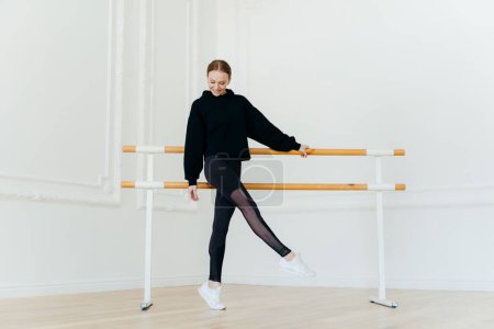 Photo for Young ballet dancer has warm up, stands near handrails, dressed in black sportclothes, stands on tip toe, wears white sneakers, focused down on floor, has cheerful facial expression, raises leg - Royalty Free Image